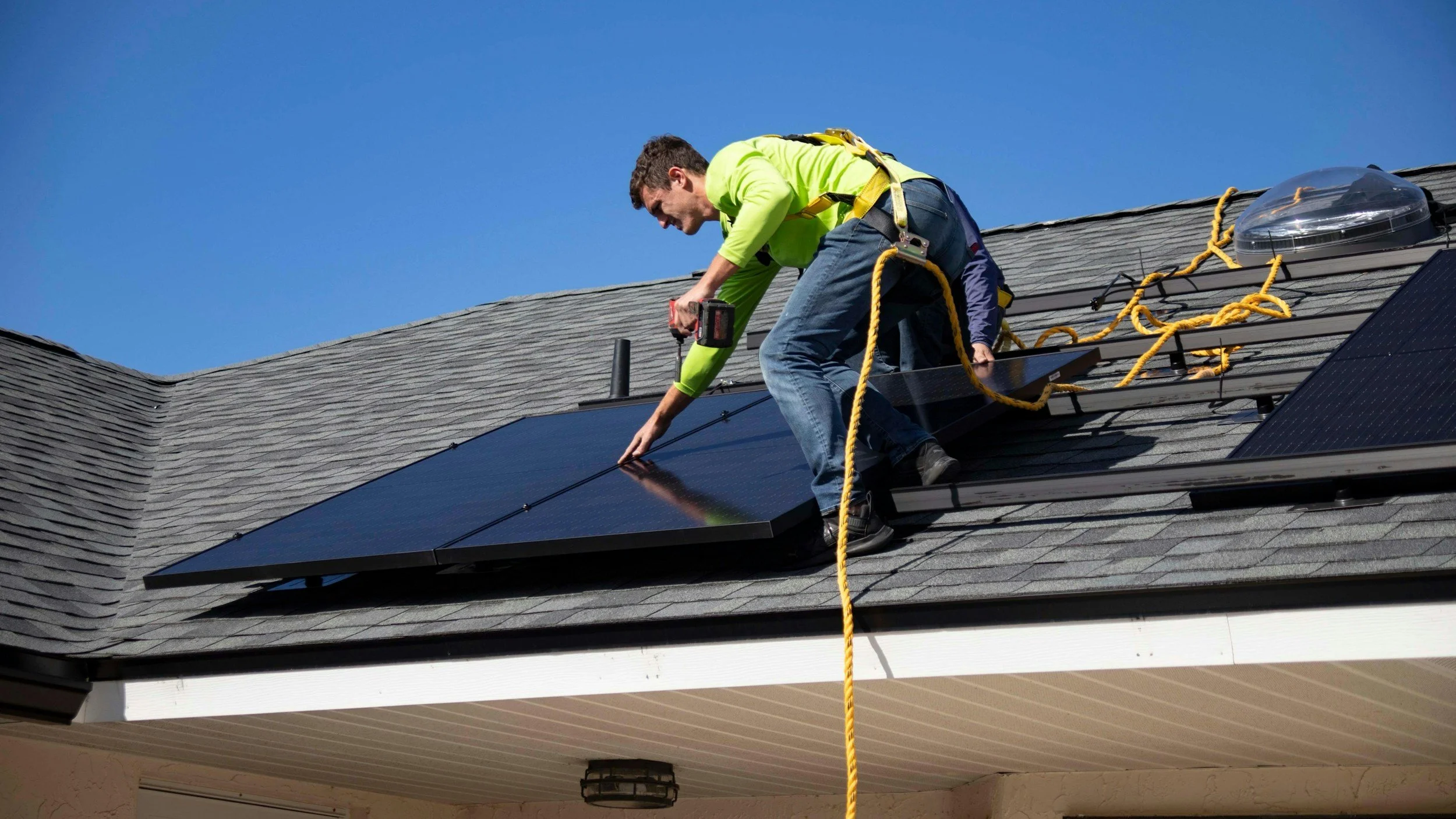 A man installing solar panels on a house roof under a clear blue sky.