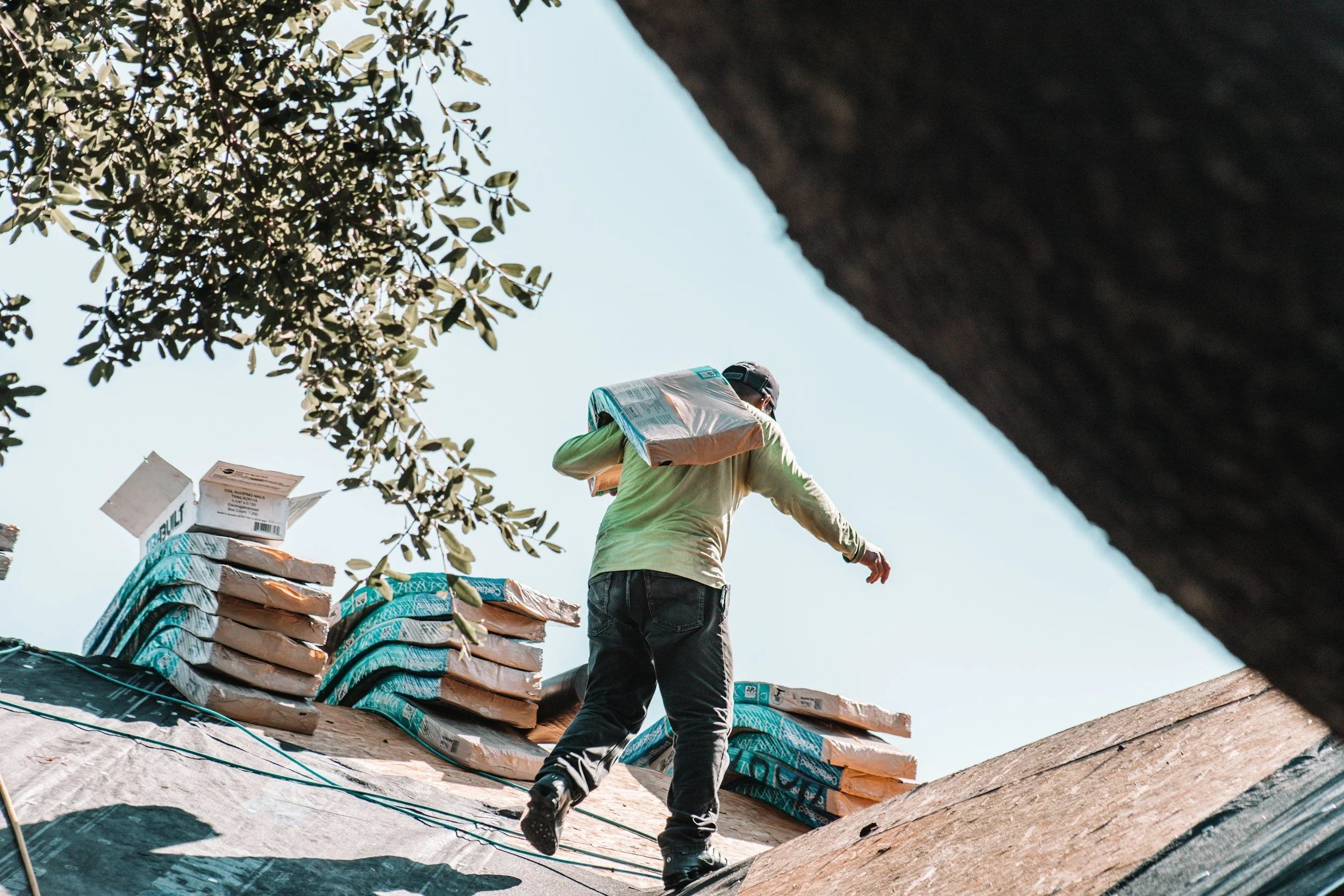 A construction worker carrying roofing materials on his shoulder on a roof under a clear blue sky. Piles of roofing shingles are visible nearby.