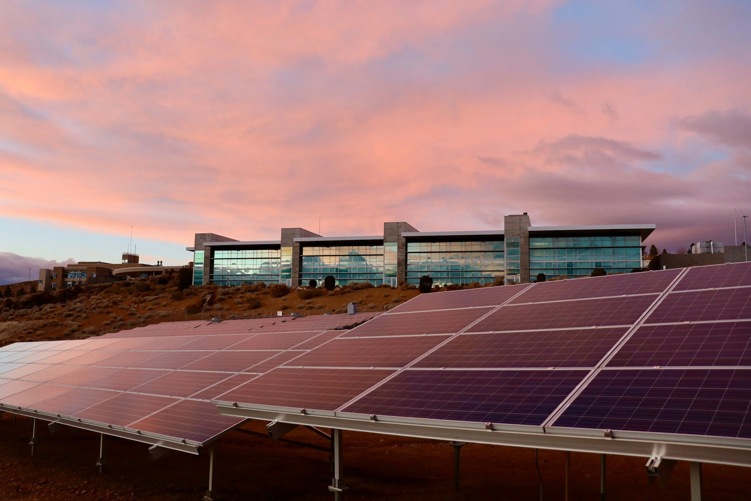 Solar panels on a hillside with a modern building and a pink and purple sunset sky in the background.