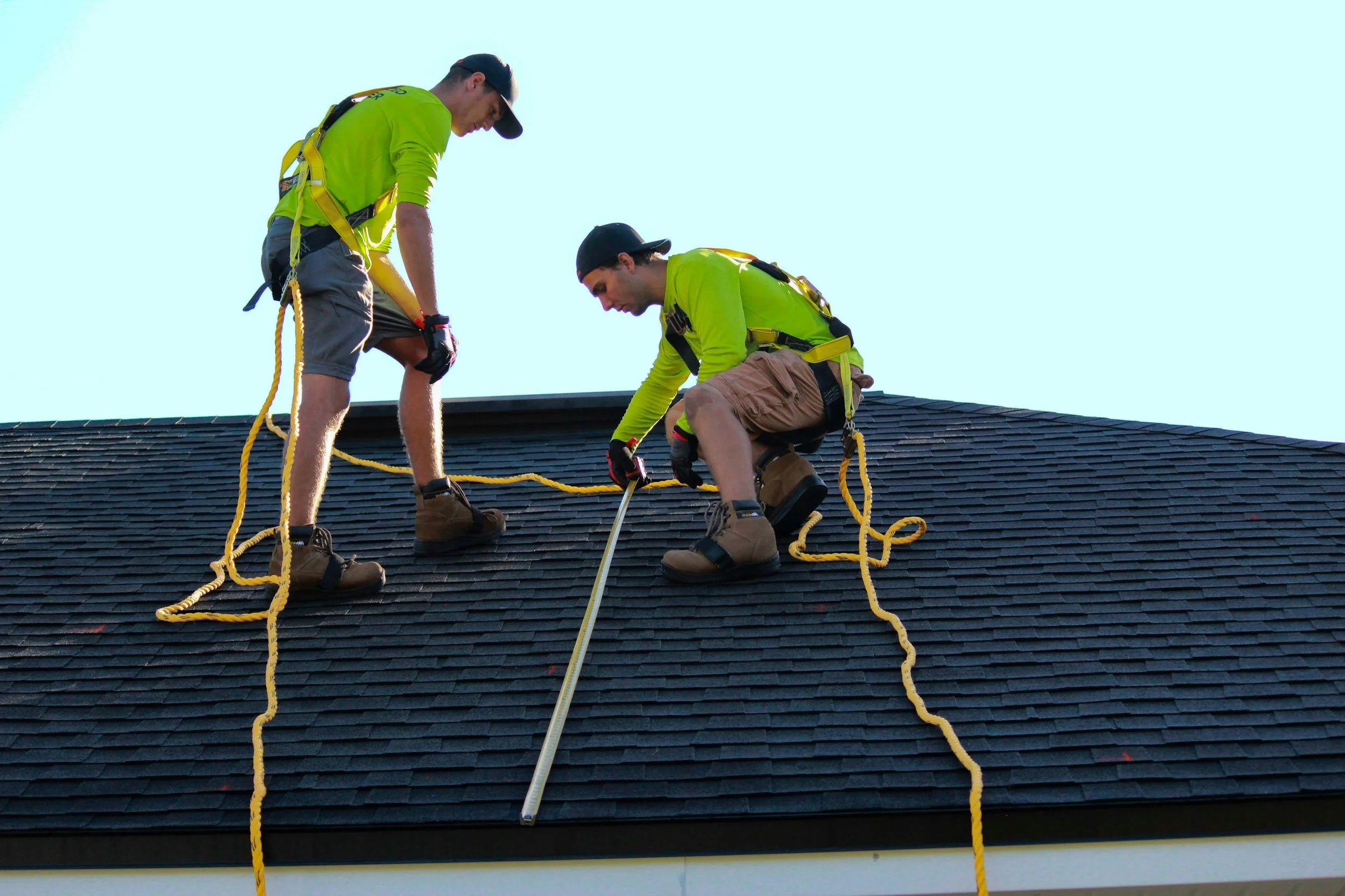 Two construction workers working on a roof, wearing safety harnesses and gloves, one handling a measuring tool.